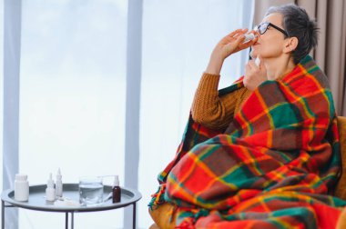 Soğuk algınlığı ya da nezle olan yaşlı bir kadın, burun spreyi sıkarak, evde battaniyeye sarılmış, yardım arıyor.