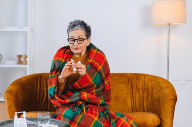 Yaşlı kadın grip belirtileriyle kendini iyi hissetmiyor. Kanepeye sarılıp burun spreyi sıkıyor.