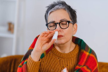 Soğuk ya da alerji sezonunda evde burun spreyi kullanan, battaniyeye sarılı yetişkin bir kadın.