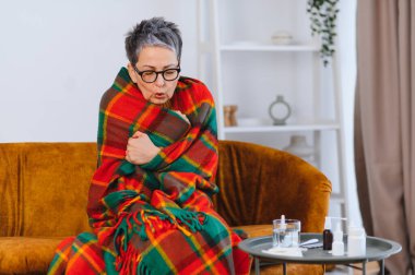 Kendini hasta hisseden bir kadın, sıcak bir battaniyeye sarılıp evdeki kanepede soğuktan titriyor.