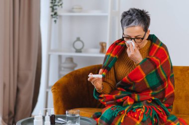 Son sınıf öğrencisi bir kadın, evde kendini kötü hissederken burnunu mendille silip ateşini kontrol ediyor.