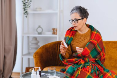 Yaşlı kadın soğuk semptomlarla savaşıyor, elinde bir burun spreyi ve battaniyeli bir kanepede mendil tutuyor, yardım arıyor.