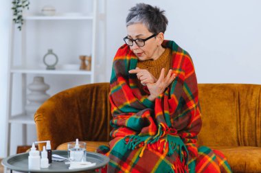 Yaşlı kadın battaniyeye sarılmış, soğuk algınlığı ya da gripten titriyor, yanında ilaçlarla kanepede dinleniyor.