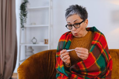 Yaşlı kadın kendini iyi hissetmiyor, battaniyeye sarılıp ateşini ölçüyor ve yüksek ateşe tepki veriyor.