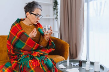 Kendini kötü hisseden bir kadın, kanepeye sarılmış, bir bardak suyla ilaç almaya hazırlanıyor.