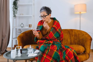 Üst düzey bir kadın, battaniyeye sarılmış, termometreyi tutuyor, hastalığa kafa yoruyor ve ilaç alıyor.