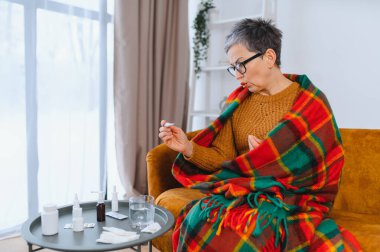 Soğuk algınlığı ve grip belirtileri gösteren son sınıf öğrencisi bir kadın kanepeye sarılıp battaniyeye sarılı bir termometreyle ateşini ölçüyor.