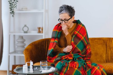 Olgun bir kadın öksürürken ağzını kapatıyor, hasta hissediyor ve evinde sıcak bir battaniyeye sarılıyor. İlaçlar masada.