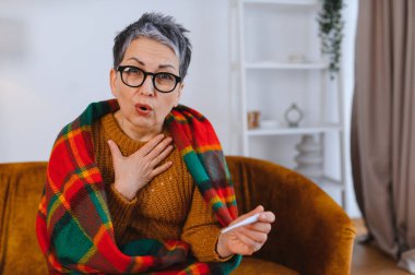 Yaşlı kadın kendini iyi hissetmiyor, battaniyeye sarılmış, termometreyi tutuyor ve grip belirtileri gösteriyor.