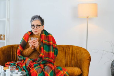 Yaşlı kadın evde dinleniyor, sıcak bir battaniyeye sarılmış kanepede, sıcak çayını yudumluyor ve soğuk algınlığından veya gripten dinleniyor.