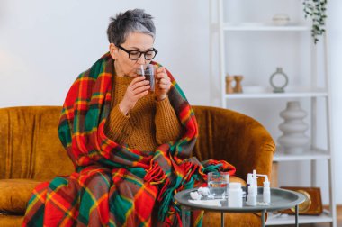 Soğuk algınlığı ya da gripten muzdarip yaşlı bir kadın battaniyeye sarılıp sıcak çay içiyor.