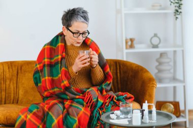 Kanepede rahat bir battaniyeye sarılı hastalıktan muzdarip yaşlı bir kadın, çay içiyor, iyileşmek için ilaçlarını yakınlarda içiyor.