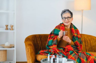 Yaşlı kadın kendini kötü hissediyor, kanepede oturuyor, ekose bir battaniyenin altında ısınıyor, iyileşmek ve rahatlamak için sıcak çay içiyor.