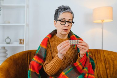Yaşlı kadın, sıcak bir battaniyeye sarılı bir kanepede otururken elinde ilaç haplarıyla kendini kötü hissediyor.