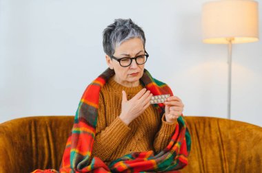 Yaşlı kadın battaniyeye sarılmış, grip ya da soğuk algınlığı semptomlarından rahatsız, elinde bir paket hap tutuyor.