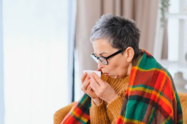 Yaşlı kadın grip belirtilerinden rahatsız, sıcak bir battaniyeye sarılmış halde burnunu beyaz bir dokuya sümkürüyor.