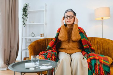 Yaşlı bir kadın şakaklarını sıkıca tutuyor. Kanepede otururken şiddetli migren ağrısı çekiyor.