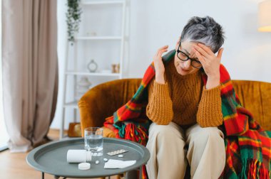 Kanepede oturan olgun bir kadın şakaklarını tutarken migren ağrısı çekiyor. İlaçlar ve bir bardak su masanın üstünde.