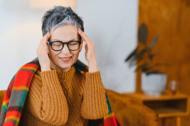 Şiddetli migrenden muzdarip yaşlı bir kadın, gözleri kapalı şakaklarına dokunuyor, açıkça rahatsızlık ve stres gösteriyor.