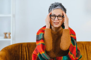 Yaşlı kadın güçlü bir baş ağrısı ya da migrenden muzdarip, kanepeye sarılmış, şakaklarına dokunuyor.