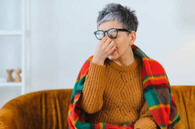 Kıdemli bir kadın burnunu kapatıyor ve kendini iyi hissetmiyor, sıcak bir battaniyeye sarılmış, nezle, grip ya da migren geçiriyor.