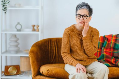 Kanepede kıdemli bir kadın yüz yüze, diş ağrısı ve diş ağrısı çekiyor, endişeli ve rahatsız.