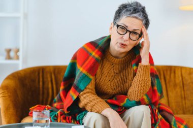 Yaşlı kadın kendini kötü hissediyor, battaniyeye sarılmış bir koltukta oturuyor, şiddetli bir baş ağrısı ya da migren ağrısı çekiyor.