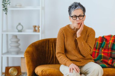 Beyaz saçlı, yaşlı bir kadın ağrılı bir diş ağrısı çekiyor, koltukta otururken yanağını tutuyor. Sağlık hizmetleri kavramı