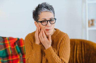 Kıdemli bir kadın diş ağrısı yüzünden yüzünü buruşturuyor ve yanağını tutuyor. Acil diş bakımı ve tedavisine ihtiyacı var.