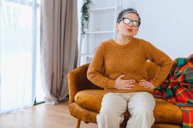 Olgun bir kadın koltukta oturup rahatsızlık hissediyor, karnını tutuyor ve hazımsızlık belirtileri gösteriyor. Sağlık ve refah kavramı