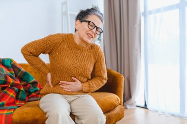 Karın ve bel ağrısından muzdarip yaşlı kadın, karnını ve yanını tutuyor, yüzünü buruşturuyor.