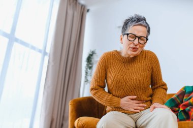 Yaşlı kadın karın ağrısı çekiyor, koltukta otururken rahatsızlık ve hazımsızlık çekiyor.