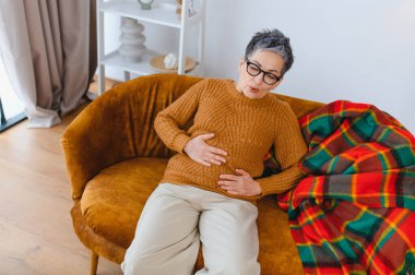 Karın ağrısı çeken bir kadın koltukta otururken, karnına ağrı saplanıyor. Sağlık, sindirim, hastalık konsepti
