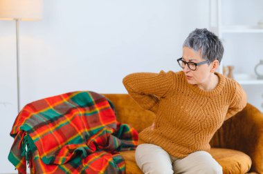 Yaşlı bir kadın evdeki koltukta otururken sırt ağrısı çekiyor, yaşlılıkta sağlık sorunlarını vurguluyor.