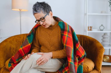 Kanepede oturan yaşlı bir kadın, ağrıyan dizini ovuyor, eklem iltihabı yüzünden rahatsızlık ve eklem ağrısı hissediyor.