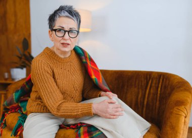 Kanepede oturan son sınıf öğrencisi kadın endişeli bir ifadeyle diz ağrısı çekiyor. Yaşlanan sağlık sorunlarını vurguluyor.