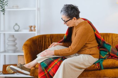 Kanepede oturan yaşlı bir kadın, eklem iltihabı veya yaralanma nedeniyle diz ağrısı ve rahatsızlık yaşıyor, bakıma ve rahatlamaya ihtiyacı var.