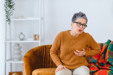 Evde kanepede oturan yaşlı bir kadın, keskin göğüs ağrısı hissediyor, potansiyel kardiyovasküler sorunları ya da kalp hastalıklarını gösteriyor.