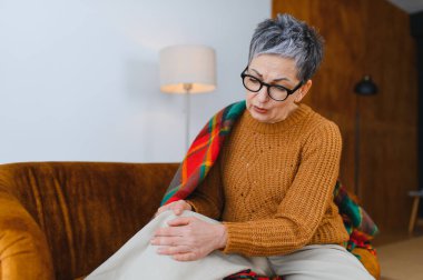 Yaşlı bir kadın dizine dokunuyor, evdeki kanepede otururken rahatsızlık ya da sakatlık geçiriyor. Yaşlılıkta sağlık sorunları