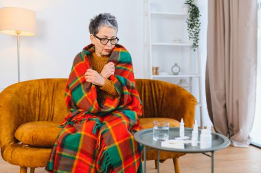 Yaşlı kadın kendini iyi hissetmiyor, sıcak bir battaniyeye sarılmış, oturma odasında grip belirtileri gösteriyor.