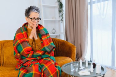 Yaşlı kadın kendini hasta hissediyor, battaniyeye sarılı bir koltukta oturuyor, grip belirtileri gösteriyor.
