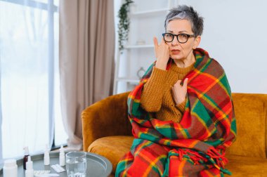 Yaşlı yetişkin bir kadın sıcak battaniyeye sarılı olarak koltukta oturuyor. Gripten rahatsız oluyor ve evde titriyor.