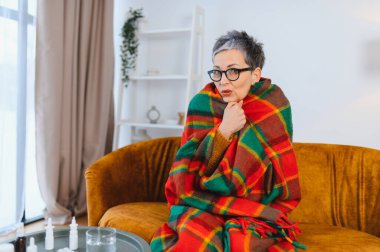 Kadın üşütmüş ya da grip olmuş, battaniyeye sarılmış, masanın üzerinde ilaçla kanepede titriyor.