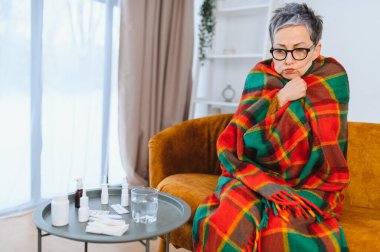 Kadın kendini iyi hissetmiyor ve titriyor. Sıcak bir battaniyeye sarılmış bir şekilde. Masasında ilaç ve suyla bir koltukta oturuyor.