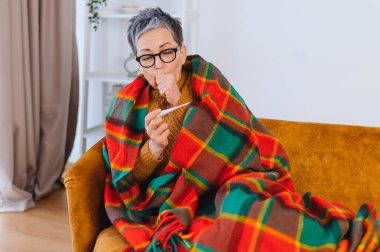Yaşlı kadın hastalıktan muzdarip, üşümüş ve öksürüyor, termometreyle ısıyı kontrol ediyor.