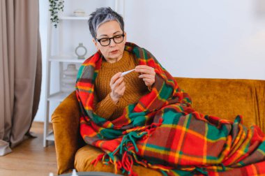 Ekoseli battaniyenin altındaki yaşlı kadın ateşini dijital termometreyle ölçüyor. Hastalık, nezle ve grip kavramları