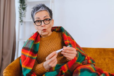 Yaşlı kadın kendini hasta hissediyor, battaniyeye sarılmış, ateşini ölçüyor ve şaşırmış bir ifade sergiliyor.