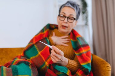 Yaşlı kadın kendini iyi hissetmiyor, sıcak bir battaniyeye sarılmış ve sıcaklığını dijital termometreyle ölçüyor.