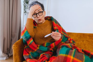 Yaşlı bir kadın soğuk algınlığı ya da gripten dolayı kendini iyi hissetmiyor, termometreyle ölçüyor, kanepede sıcak bir battaniyeyle örtüyor.