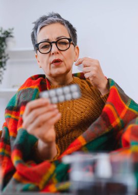 Sıcak bir battaniyeye sarılmış, grip ya da soğuk algınlığı geçiren, rahatlaması için bir paket hap tutan olgun bir kadın.